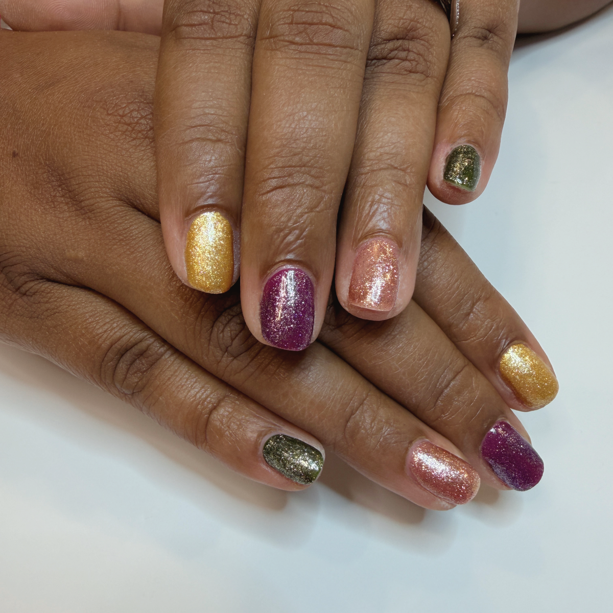 Short, rounded nails with shimmering gold, plum, rose gold, and olive green polish.