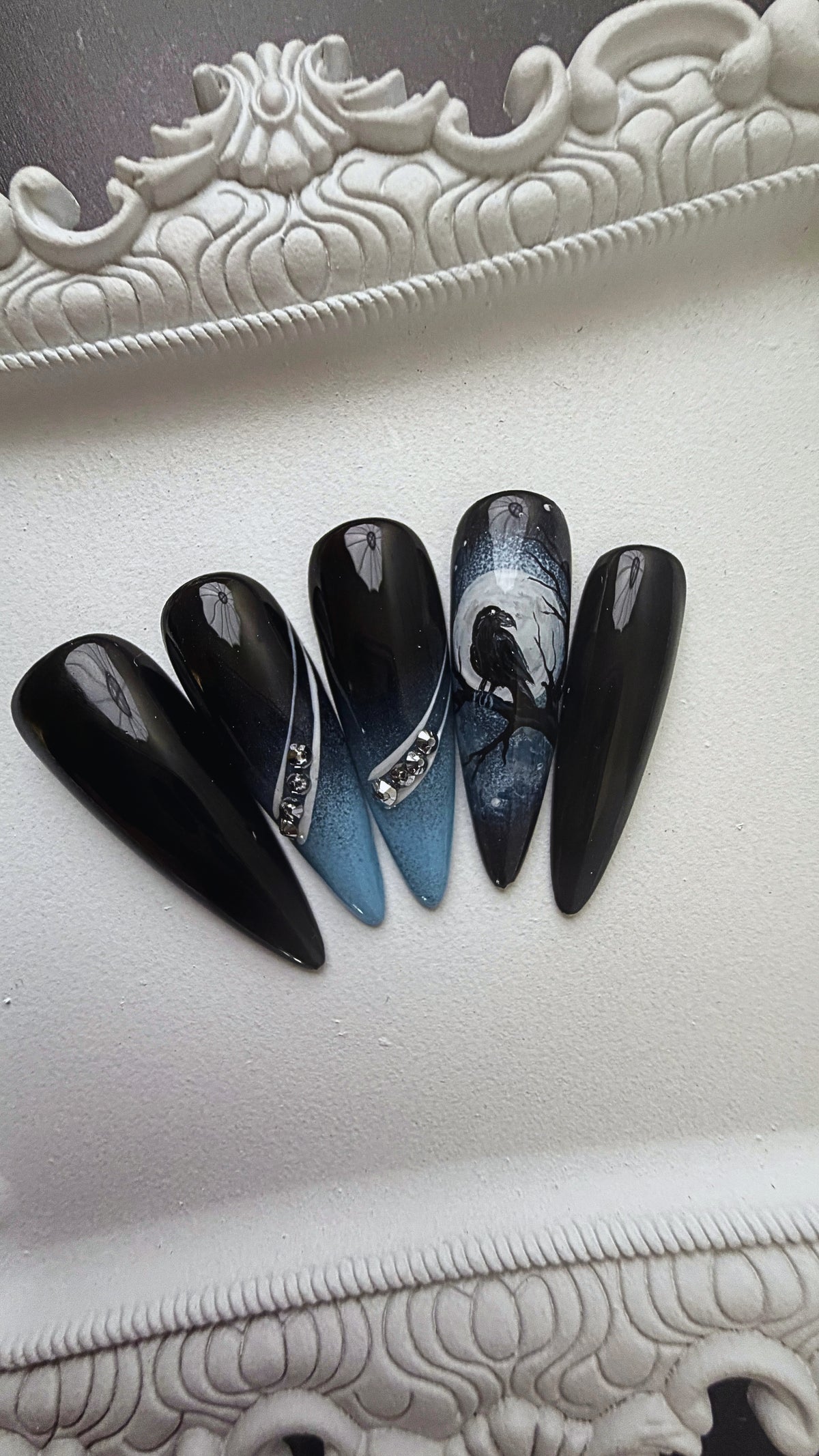 Black-and-blue stiletto press-on nails with raven artwork and crystal accents.