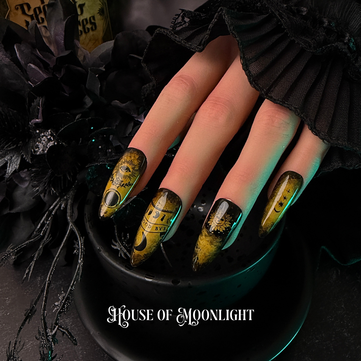 A close-up of a hand with gothic nail art featuring yellow and black celestial ouija designs, resting in a black bowl with the text 'House of Moonlight' at the bottom.