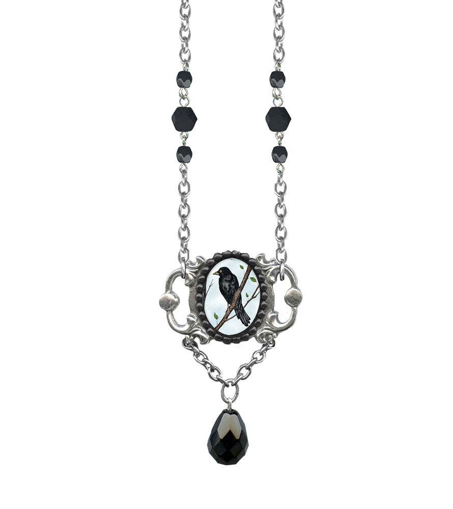 A gothic-style silver chain necklace featuring black faceted beads, an ornate oval pendant with a black bird on a branch over a white mother-of-pearl background, and a dangling faceted black teardrop charm, shown against a plain white background.